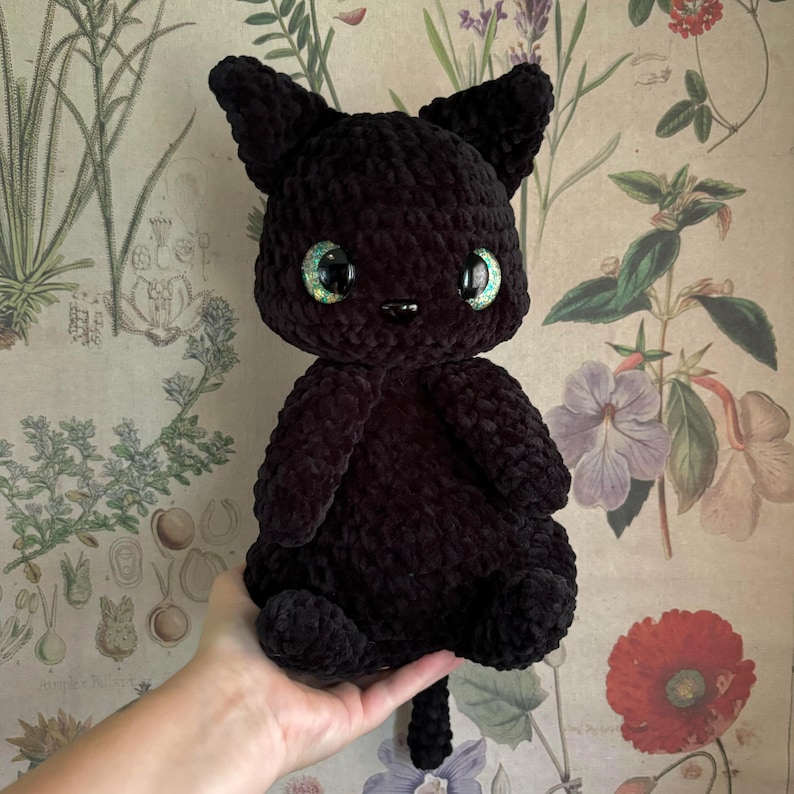 May include: A black crocheted cat plush toy with green eyes. The cat is sitting on a hand and has a long tail.