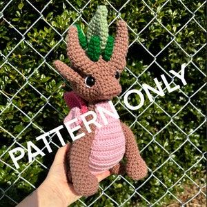 May include: A crocheted brown dragon with a pink belly and green leaves on its head. The dragon is being held in a hand. The background is a chain link fence.