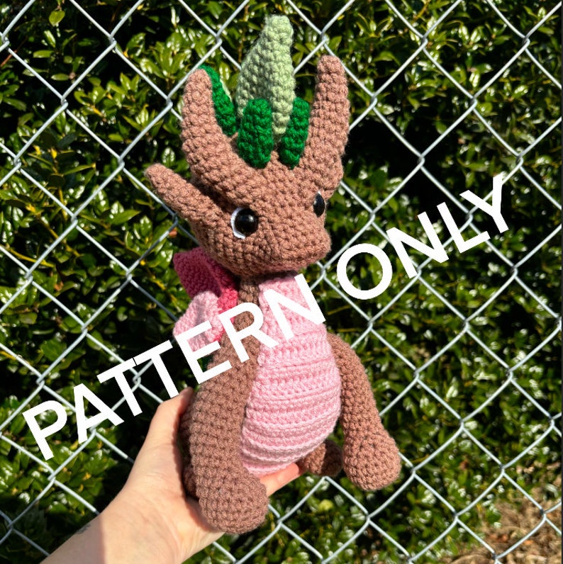 May include: A crocheted brown dragon with a pink belly and green leaves on its head. The dragon is being held in a hand. The background is a chain link fence.