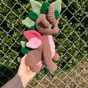 May include: A brown crocheted dragon with green and pink accents. The dragon has a spiky back, wings, and a horn on its head.