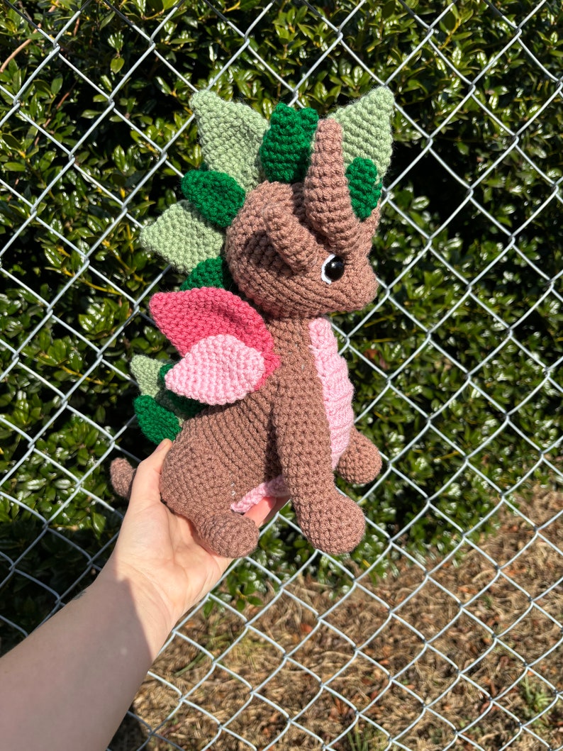 May include: A brown crocheted dragon with green and pink accents. The dragon has a spiky back, wings, and a horn on its head.