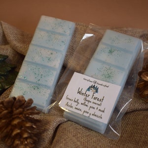 May include: Two light blue wax melt bars with green glitter, packaged in clear plastic. The label reads "Winter Forest" with scent notes. A pine cone, Christmas tree, and gift box are in the background.