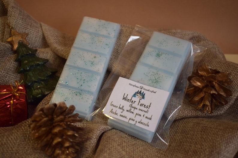 May include: Two light blue wax melt bars with green glitter, packaged in clear plastic. The label reads "Winter Forest" with scent notes. A pine cone, Christmas tree, and gift box are in the background.
