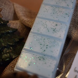 May include: A light blue wax melt bar with green glitter, next to a green Christmas tree ornament and a gold star. The items are arranged on a burlap surface, suggesting a holiday theme.
