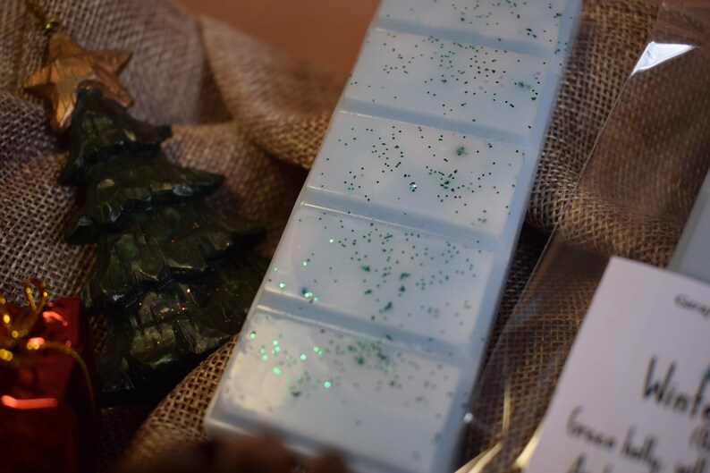 May include: A light blue wax melt bar with green glitter, next to a green Christmas tree ornament and a gold star. The items are arranged on a burlap surface, suggesting a holiday theme.