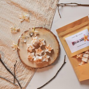 Könnte beinhalten: Eine braune Papiertüte mit einem weißen Etikett, auf dem "Flor de Tahiti" steht und ein Bild einer weißen Blume zu sehen ist. Die Tüte enthält kleine weiße Wachs-Schmelzen in Sternform. Die Wachs-Schmelzen liegen auf einer runden Holzscheibe, um die herum weiße Blüten verstreut sind.