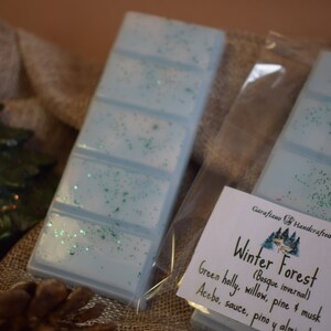 May include: Two light blue wax melt bars with green glitter, packaged in clear plastic. The label reads "Winter Forest" with scent notes. Festive decorations, including a Christmas tree and star, are in the background.