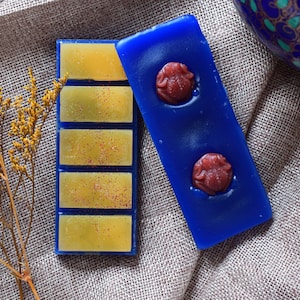 May include: Two wax melts, one blue with two red wax embellishments and one yellow with a blue border and glitter.