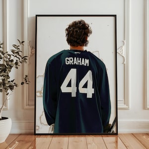 May include: Framed print featuring the back of a hockey jersey. The navy blue jersey displays the name "GRAHAM" above the number "44" in white. The person's dark hair is visible. The print is in a black frame.