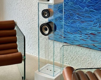 May include: A modern, clear glass speaker tower with two visible speakers. The tower has a white base and top, and is set against a textured white wall and a blue abstract painting. The speakers are black and brown.