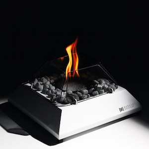 以下が含まれることがあります： ガラス製のエンクロージャーとメッシュスクリーンを備えた、モダンな白い卓上暖炉。暖炉には灰色の石と明るいオレンジ色の炎が入っています。「BERLING DESIGN」のブランド名が側面に印刷されています。