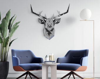May include: A white wall with a geometric silver deer head sculpture, two blue armchairs, a small white table with a book and two decorative vases, and a floor lamp with a white globe.