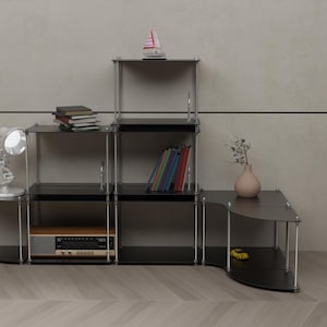 May include: A black metal shelf unit with five shelves. The shelves are filled with books, a small toy sailboat, a lamp, and a vase with flowers. The shelf unit is against a light gray wall.