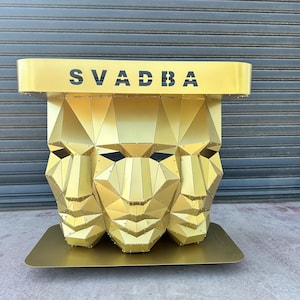 Op de afbeelding: Een goudkleurige metalen tafel met een geometrisch low-poly ontwerp van drie samengevoegde gezichten als basis. Het rechthoekige tafelblad heeft het woord SVADBA in zwarte hoofdletters op de voorrand, met een gladde, platte voetplaat.