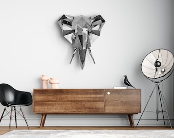 May include: A silver geometric elephant head wall sculpture hangs above a wooden dresser with two drawers. The dresser has a black chair in front of it and a black tripod lamp with a white reflector to the right.
