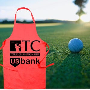 Könnte beinhalten: Eine leuchtend rote Schürze mit dem schwarzen Text "TC TOUR CHAMPIONSHIP usbank". Die Schürze befindet sich auf einem grünen Golfplatz mit einem blauen Golfball im Hintergrund. Die Schürze hat eine Nackenschlaufe und Bindebänder.