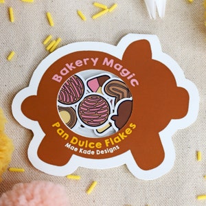 May include: A brown and white pig-shaped sticker sheet with a clear window showing colorful stickers of Mexican pastries. The text on the sticker sheet reads "Bakery Magic Pan Dulce Flakes Mae Kade Designs".