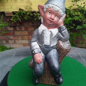 May include: A grey and white garden gnome with a pointed hat sits on a tree stump. The gnome has a white collar and is wearing a grey outfit. The gnome is looking to the side with a thoughtful expression.