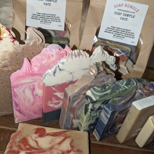 May include: A variety pack of handmade soap bars in different colors and patterns. The soap bars are arranged in a display on a wooden surface. The soap bars are made with shea butter, cocoa butter, avocado oil, coconut oil, and olive oil. The soap bars are labeled "Soap Sample Pack".