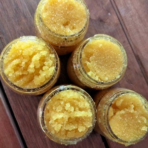 May include: Five small glass jars filled with a yellow-gold sugar scrub. The jars are arranged in a pyramid shape on a wooden surface.