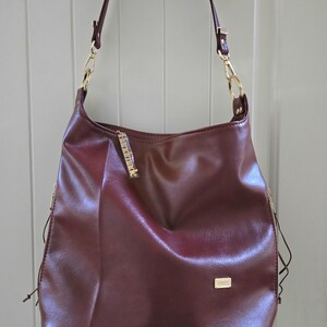 May include: A burgundy shoulder bag with gold-colored chain straps and accents. The bag has a rectangular gold-colored tag and a small gold-colored charm. The bag is hanging from a silver-colored metal bar.