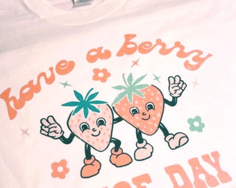 May include: White t-shirt with a retro design featuring two smiling strawberry characters. The text "have a berry NICE DAY" is printed in peach-colored letters. The shirt has a crew neck and short sleeves.