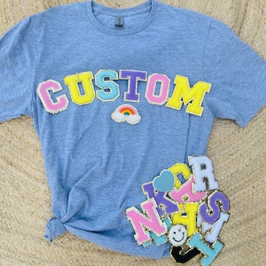 May include: A light blue t-shirt with the word "CUSTOM" in colorful felt letters. The letters are outlined in gold glitter. There are other colorful felt letters and patches on the shirt, including a rainbow.