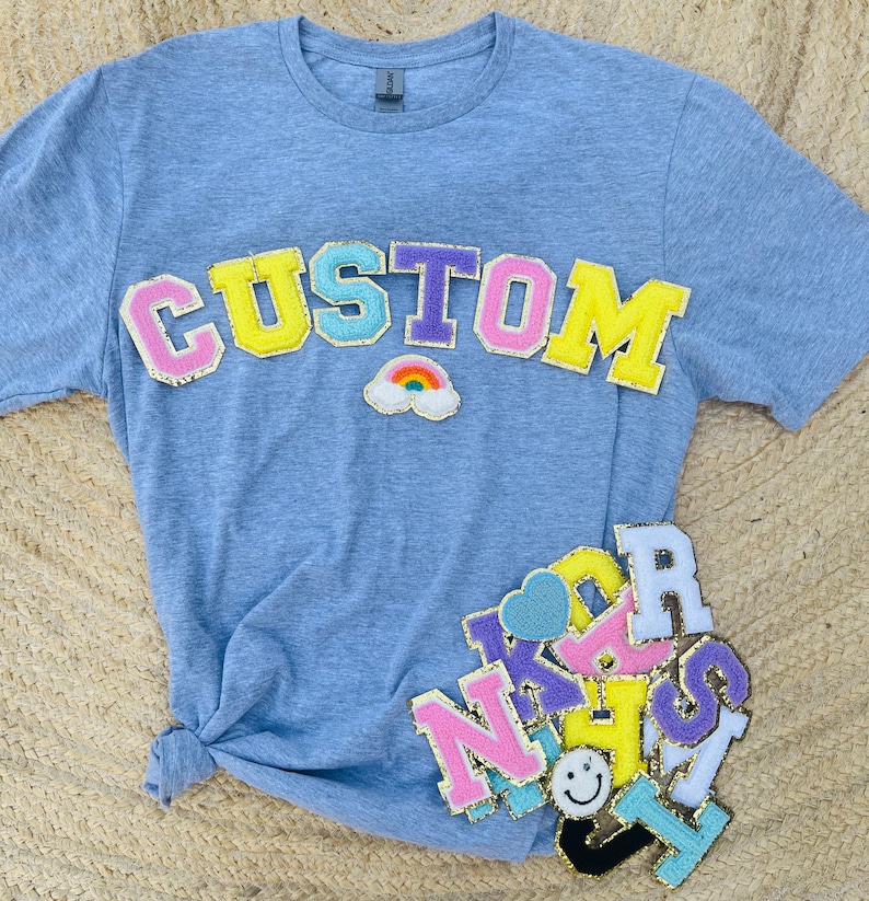 May include: A light blue t-shirt with the word "CUSTOM" in colorful felt letters. The letters are outlined in gold glitter. There are other colorful felt letters and patches on the shirt, including a rainbow.