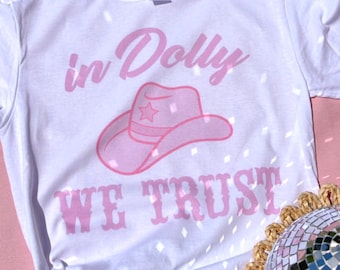 May include: White t-shirt with pink text that reads "in Dolly we trust" and a pink cowboy hat with a star on it.
