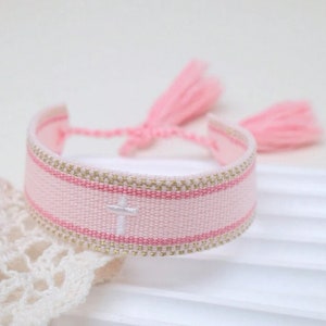 May include: A pink woven bracelet with gold trim and a white cross. The bracelet has pink tassels and a pink and gold striped design. The bracelet is a religious accessory.