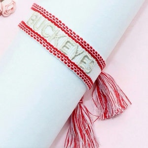 May include: White and red woven bracelet with the word "BUCKEYES" embroidered in white. The bracelet has red and white tassels.