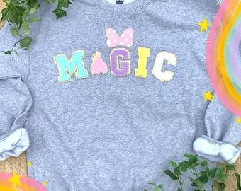 May include: A gray sweatshirt with the word "MAGIC" in colorful, glittery letters. A pink bow with white polka dots is above the word. The sweatshirt is on a light brown background with a rainbow and stars.