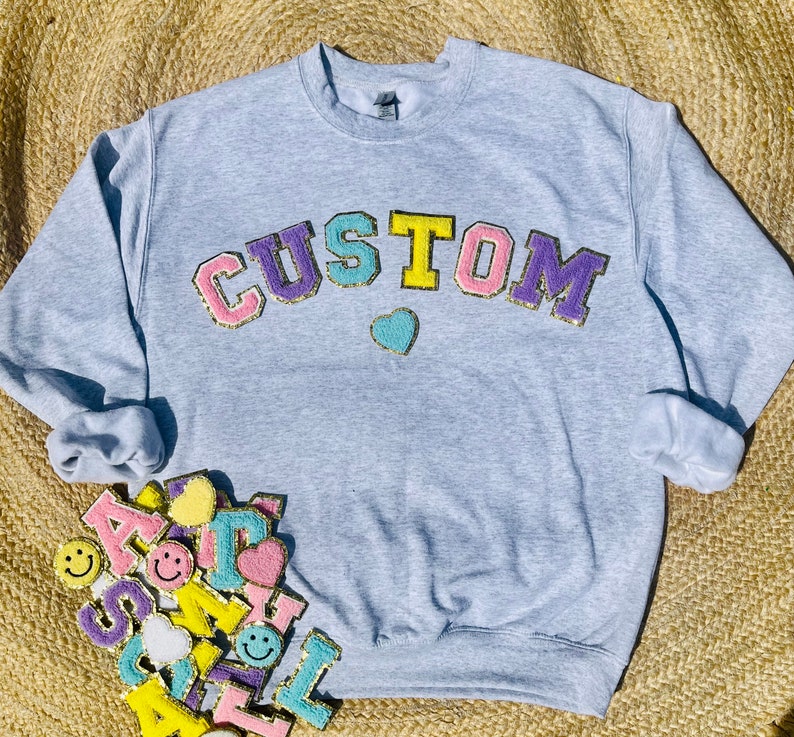 May include: A light gray sweatshirt with the word "CUSTOM" in colorful, fuzzy letters. The letters are outlined in gold glitter. A small, light blue heart is below the word.