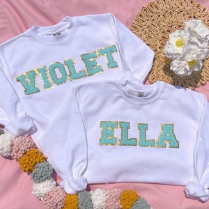 May include: Two white long-sleeved sweatshirts with gold and light blue lettering. One sweatshirt says "VIOLET" and the other says "ELLA".