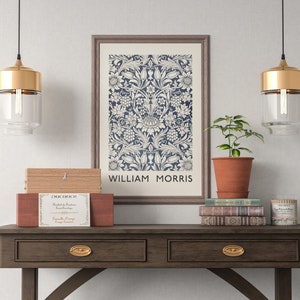 May include: A framed print of a floral pattern in blue and white with the text "William Morris" at the bottom. The print is hanging on a white wall above a wooden console table with two drawers. There are two gold pendant lights hanging above the table, a potted plant, and a stack of books on the table.