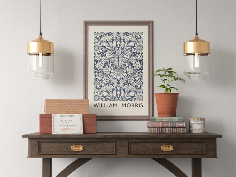 May include: A framed print of a floral pattern in blue and white with the text "William Morris" at the bottom. The print is hanging on a white wall above a wooden console table with two drawers. There are two gold pendant lights hanging above the table, a potted plant, and a stack of books on the table.