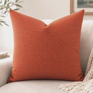 May include: A square throw pillow in a vibrant burnt orange color. The pillow features a distinct, uniform waffle-knit or basket-weave texture across its entire surface, providing a tactile, raised grid pattern that covers the front of the cushion.