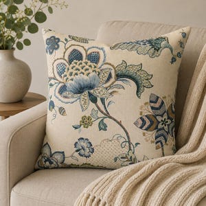 May include: A square throw pillow featuring a cream-colored base with a detailed woven floral pattern in shades of blue, green, and tan. The design includes large stylized blossoms, scrolling vines, and intricate leaf motifs across the fabric.