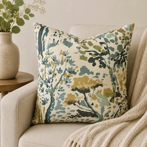 May include: A square throw pillow featuring an abstract painterly print of trees and foliage in shades of forest green, teal, mustard yellow, and dark charcoal on a cream-colored fabric background, showcasing a textured weave and piped edges.