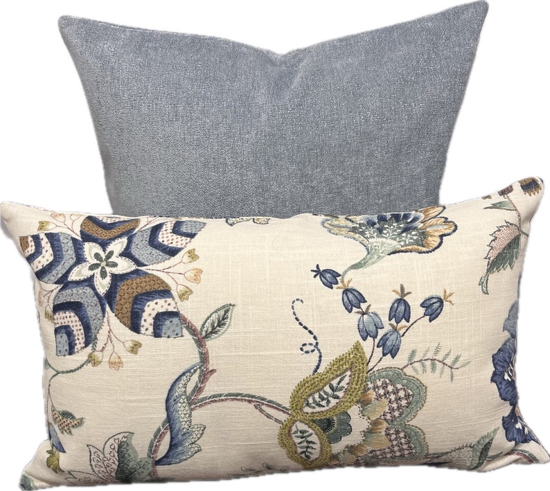 Puede incluir: Dos almohadas decorativas, una con un patr&oacute;n floral en tonos azules, verdes y marrones sobre un fondo crema, y la otra de color azul claro s&oacute;lido.