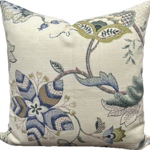 Puede incluir: Una almohada de color crema con un patr&oacute;n floral en tonos azules, verdes y marrones. La almohada presenta una gran flor estilizada en el centro, rodeada de flores y hojas m&aacute;s peque&ntilde;as.