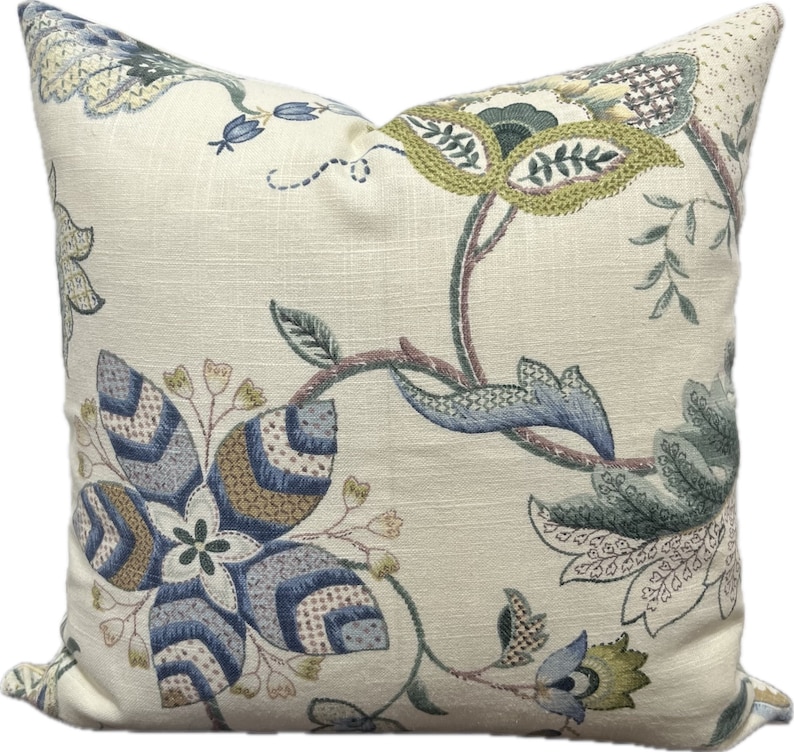Puede incluir: Una almohada de color crema con un patr&oacute;n floral en tonos azules, verdes y marrones. La almohada presenta una gran flor estilizada en el centro, rodeada de flores y hojas m&aacute;s peque&ntilde;as.