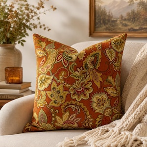 May include: A square throw pillow featuring a vibrant burnt orange base color with an intricate, multi-colored botanical and paisley pattern in shades of olive green, mustard yellow, cream, and deep red, finished with a clean, piped edge.