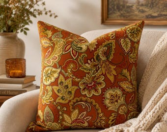May include: A square throw pillow featuring a vibrant burnt orange base color with an intricate, multi-colored botanical and paisley pattern in shades of olive green, mustard yellow, cream, and deep red, finished with a clean, piped edge.