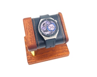 May include: A silver smartwatch with a black leather band is displayed on a wooden watch stand. The watch face has a complex design with multiple hands and a date display. The stand is made of dark wood with a light wood base.
