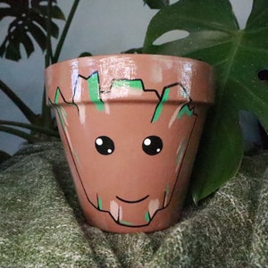 May include: A terracotta flower pot painted with a cartoon face of the character Groot from the Guardians of the Galaxy movie franchise. The pot is painted in shades of brown, green, and white.