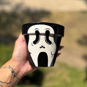May include: Black and white painted ceramic flower pot featuring a Scream mask design. The pot has a glossy black finish with a white mask graphic on the front. The pot is designed for planting flowers or other small plants.