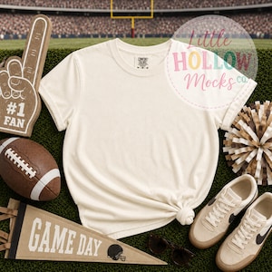 May include: A cream-colored short-sleeve t-shirt with a crew neckline. The shirt is displayed alongside a brown foam finger with #1 FAN text, a brown football, a tan pennant with GAME DAY text, a brown and white pom-pom, and beige sneakers.