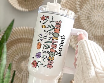 May include: White stainless steel tumbler with a handle and a straw. The tumbler has a colorful floral design and the text "Occupational Therapist" in rainbow colors.