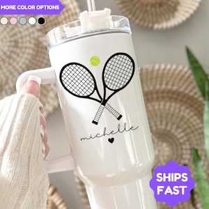 May include: White insulated tumbler featuring a tennis racket and ball design. The tumbler has a black graphic of crossed tennis rackets, a green tennis ball, and the name "michelle" in script. The tumbler has a handle and a clear lid.